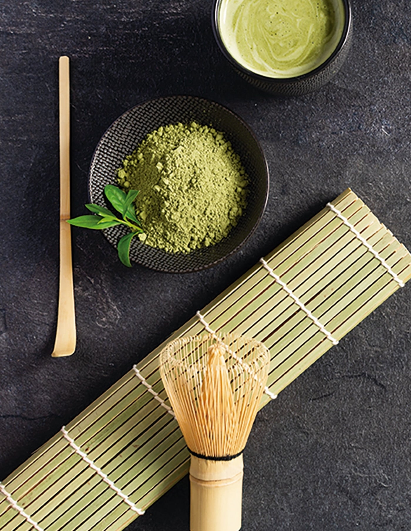 Matcha green tea powder in a bowl with a whisk and a cup of matcha on a dark surface.