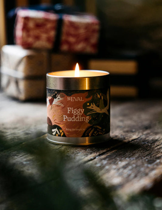 Candle with 'Figgy Pudding' label on a wooden surface, with wrapped gifts in the background.