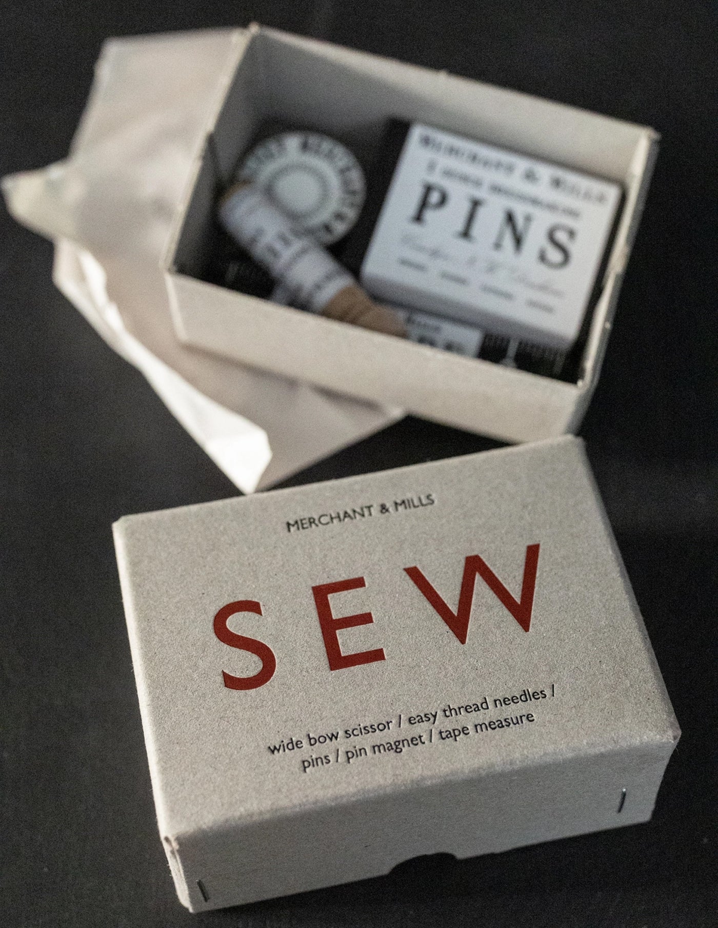 A sewing kit set in a textured light gray box with the brand name 'MERCHANT & MILLS' visible. The front box features the word 'SEW' in large red letters, and the back box is open, showing contents including a spool of thread, a tape measure, and a box of pins.